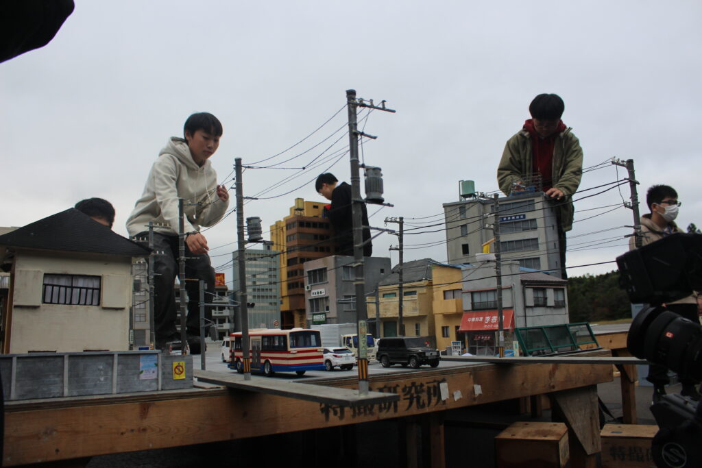 ミニチュア都市模型の上で数人が建物や電柱に手を添えて配置や演出の動きを丁寧に調整する様子の写真