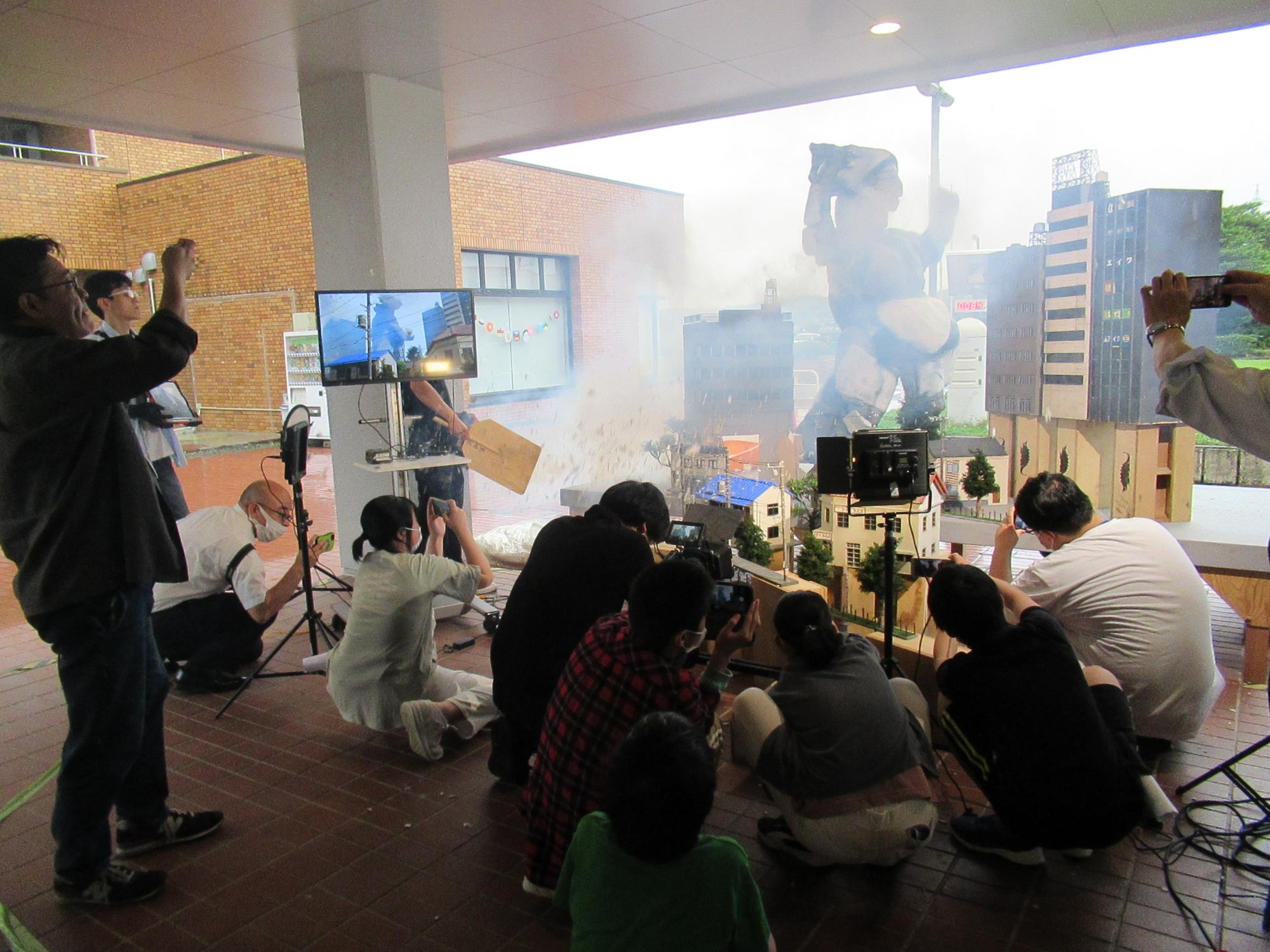 すかがわ特撮塾の活動で模擬撮影をしている様子、怪獣が中央で暴れている様子を特撮撮影しており、スモークをたくなどしている、周りで多くの人がスマートフォンを構えて撮影している