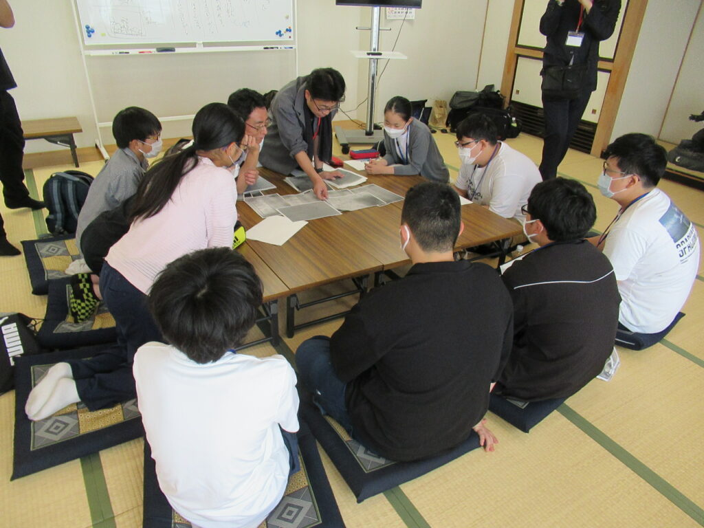 和室で長机を4つつけ大きなテーブルとし、資料を中央に配置して講師と塾生たちが取り囲んでいる様子の写真