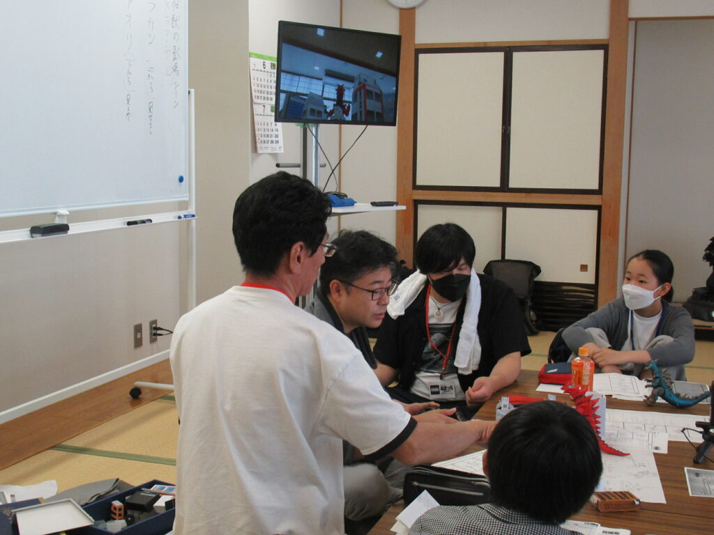 眼鏡をかけた男性講師がフィギュアや紙の資料を使い説明しているところを塾生たちが眺めている写真