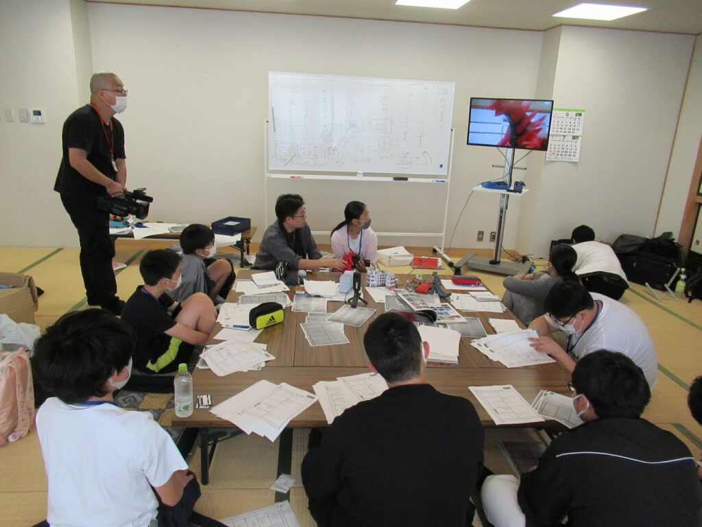 畳敷きの和室で、机を囲んで座る多くの塾生たちが資料を広げており、講師がホワイトボード横のモニターに映像を映して説明している様子の写真