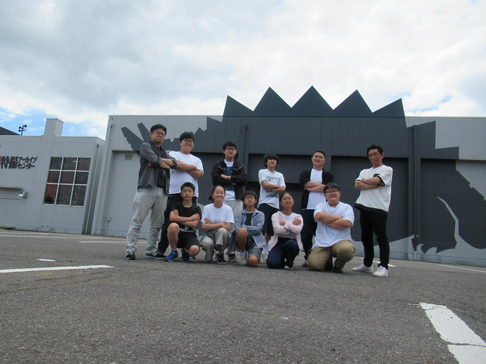 須賀川市特撮アーカイブセンターの駐車場で腕を組み屈んでいる列と立っている列の2段構成で集まっている塾生と講師の写真