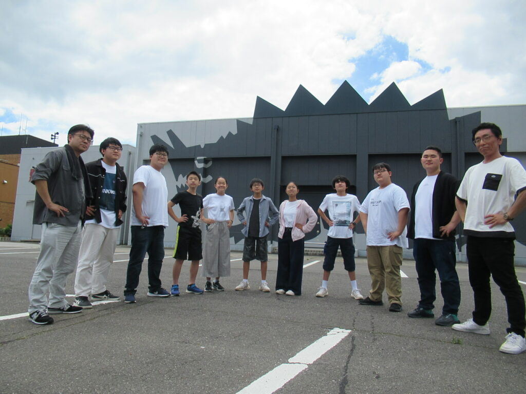 須賀川特撮アーカイブセンターの駐車場で、腰に手を置くポーズで中央に塾生、両端に講師が並んでいる写真