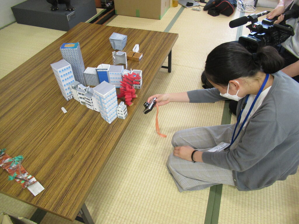 木目調の机の上で紙製の小さなビルをたくさん並べ、中央に怪獣のフィギュアを置き斜め下から写真を撮る塾生の写真