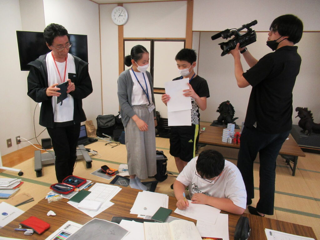 木目調の机に向かい資料に書き込みをする塾生と、資料を持ち打ち合わせをする塾生たちを見守りカメラを構える講師やスタッフたちの写真
