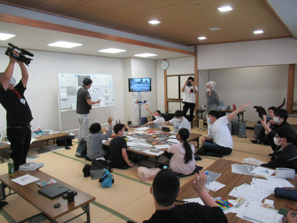 和室で長机を囲み、ホワイトボードの前に立つ講師からの質問に手を挙げる塾生たちと、その様子をカメラに収めるスタッフたちの写真