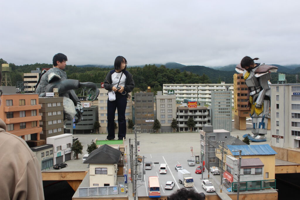 ミニチュア都市模型の上で怪獣スーツを着た人物と別の人物が建物周辺に立ち配置や動きを確認している様子の写真