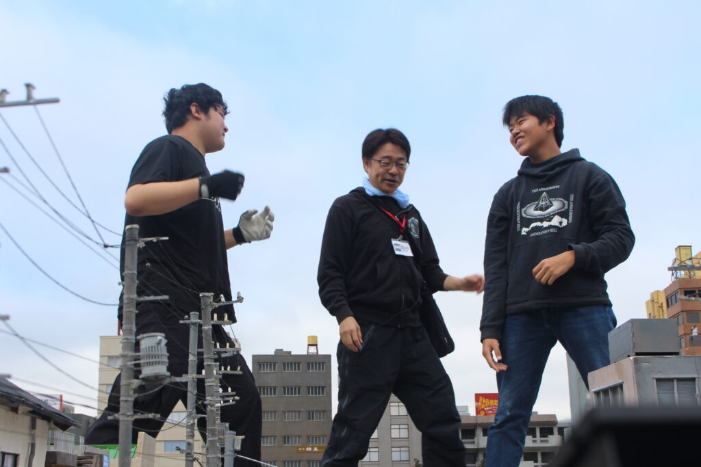 ミニチュア都市模型の上で男性3人が互いに向き合い身振りを交えて立ち位置や動作の確認を丁寧に行う様子の写真