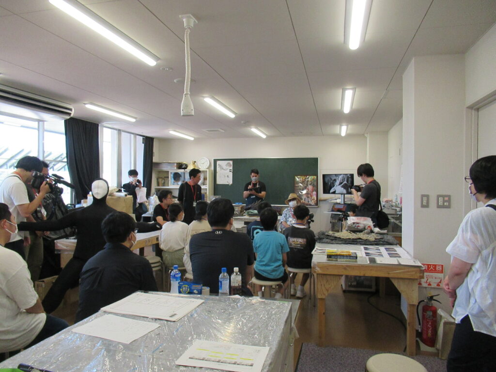 黒板の前に立つ講師の話を複数の塾生が椅子に座って真剣に聞き、その様子をカメラマンが撮影している写真