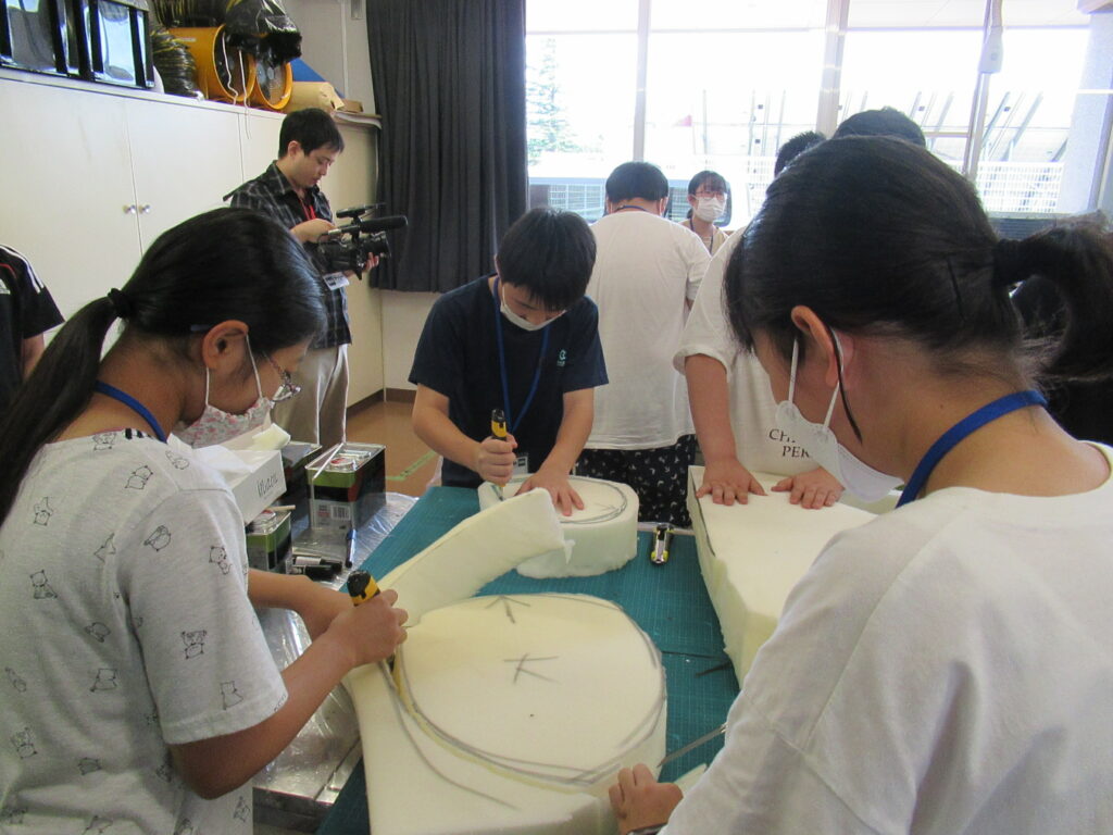 明るい教室で複数の参加者が作業台に向かい、マジックで円形の下書きが描かれた白いウレタンスポンジをカッターで慎重に切っている写真