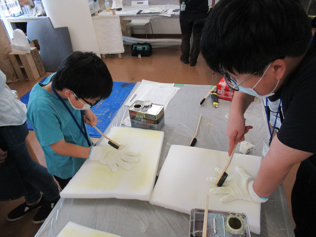 作業台の上に置かれた白いウレタンスポンジに、マスクと手袋を着用した二人が手を置きブラシを使って作業している写真