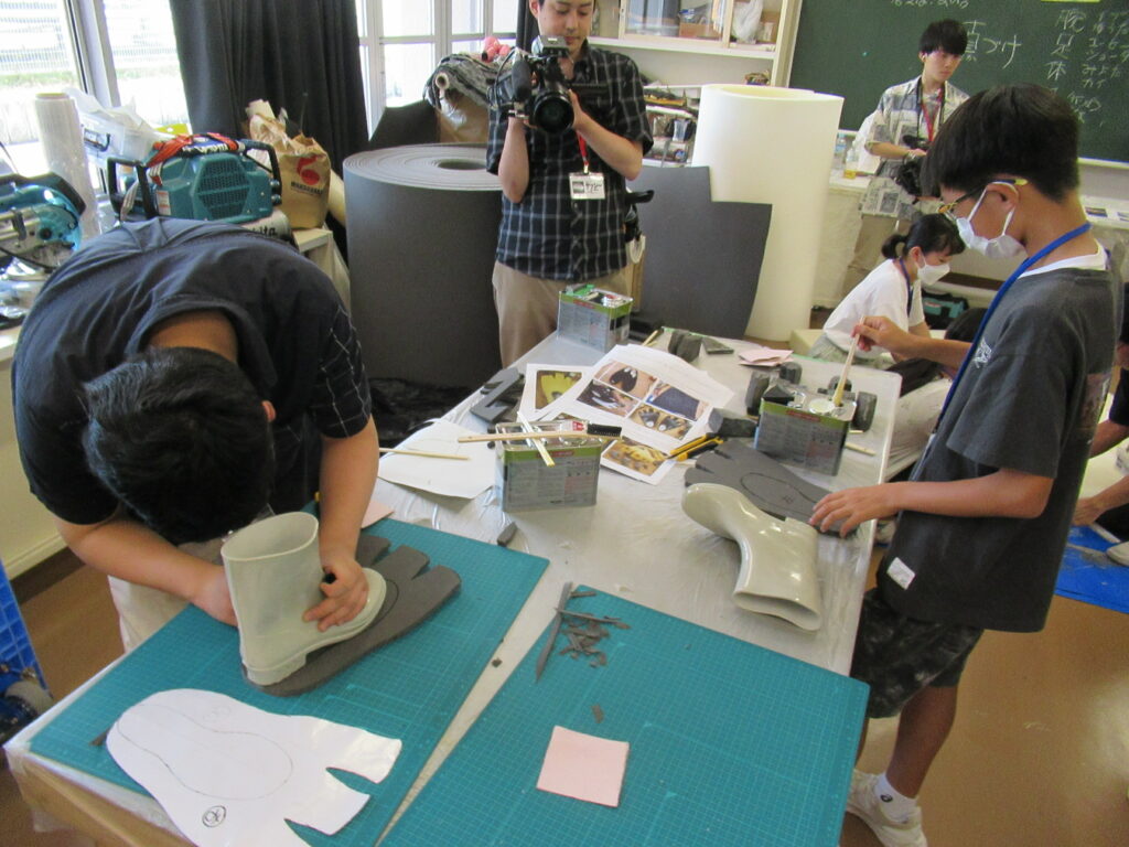 作業台の上で、足の形に切り抜いたグレーのウレタン素材に長靴を合わせたり、接着剤を準備したりしながら、怪獣の足の部分と思われるパーツを熱心に制作している写真