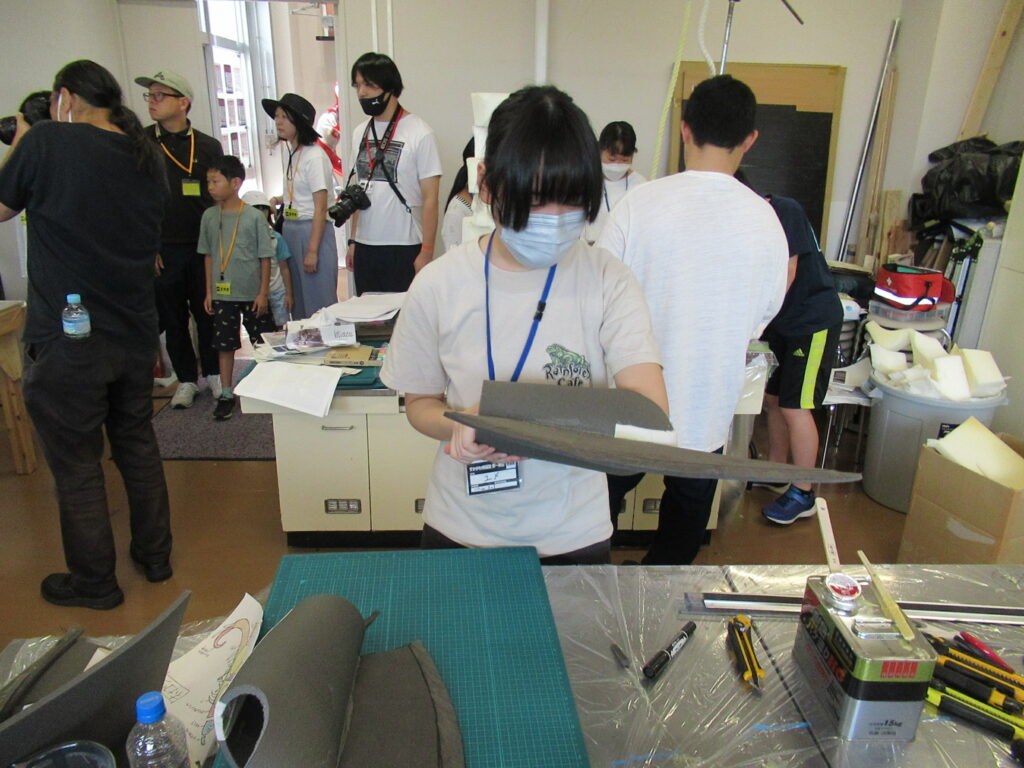 明るい室内で、マスク姿の女性参加者がグレーのウレタン素材で作られた細長い造形パーツを手に取り、仕上がりを確認している様子の写真