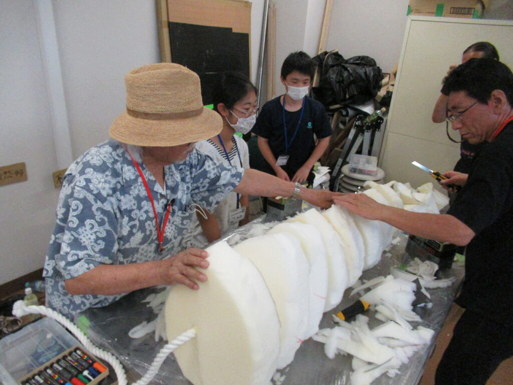 男性2人が、太いロープを通した円柱状の白いウレタンを何層も重ねて長い尻尾のような造形物を組み立てる様子を、参加者たちが真剣な表情で見守っている写真