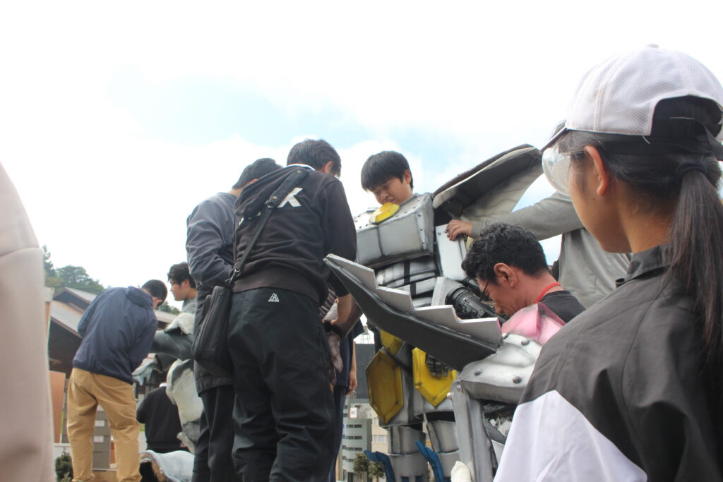 複数の人物がロボット風スーツの装着部分を囲んで調整作業を進めながら細部を確認している様子の写真