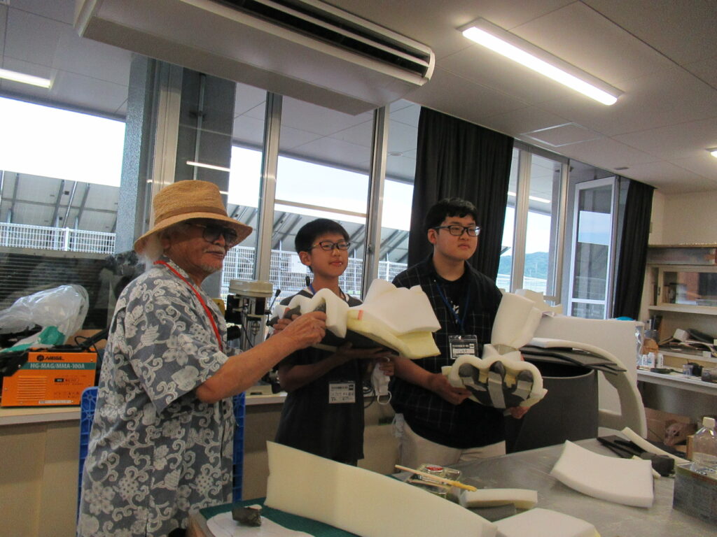 麦わら帽子を被った講師の男性と二人の少年参加者が、ウレタンスポンジで自作した怪獣の足のパーツを手に持った写真