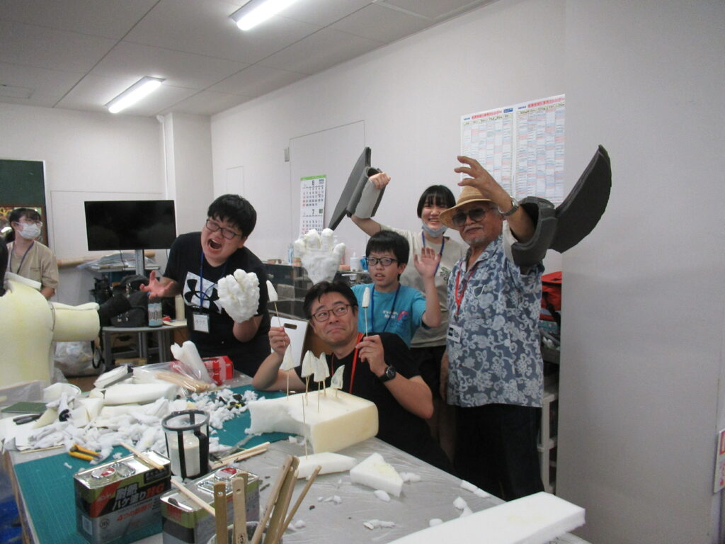 手作りの大きな白い怪獣の手や爪のパーツを装着した参加者たちが、講師の男性と共に怪獣のようなポーズをしている写真