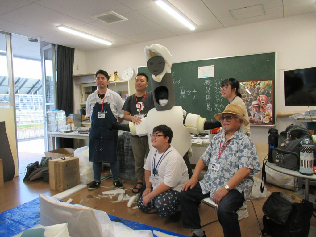 明るい教室の黒板を背にし、講師の男性とウレタン素材で自作した怪獣の着ぐるみを試着した参加者を囲んだ写真