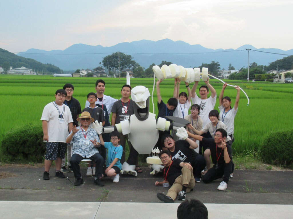 田園風景と山々を背景に、自分たちで制作した白い怪獣の着ぐるみや長い尻尾のパーツを掲げ、講師の男性を囲んで参加者がポーズを決めている写真