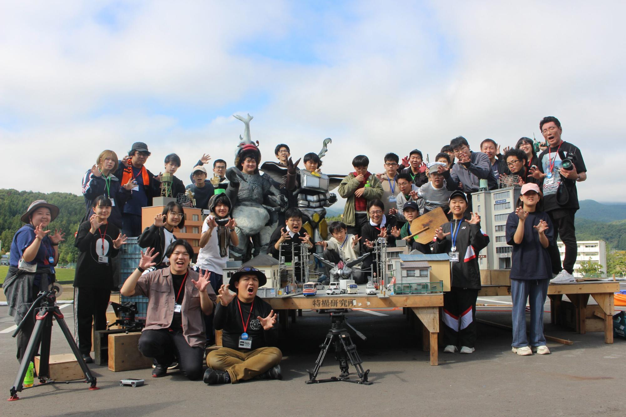 多数の参加者が巨大怪獣スーツとミニチュア都市模型を囲み揃って躍動的なポーズで記念撮影する様子の写真