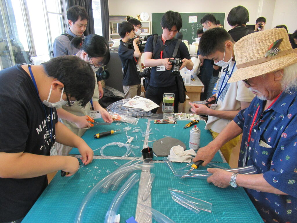 麦わら帽子を被った年配の男性と数人の若者たちが、緑色のカッティングマットが敷かれた作業台を囲み、カッターやハサミを使って透明なビニールチューブを真剣な表情で細かく裁断している写真