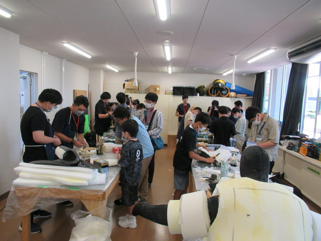 室内で多くの人々がテーブルを囲み、白い素材や道具を用いて工作や模型製作のような作業に熱心に取り組んでいる様子を捉えた写真