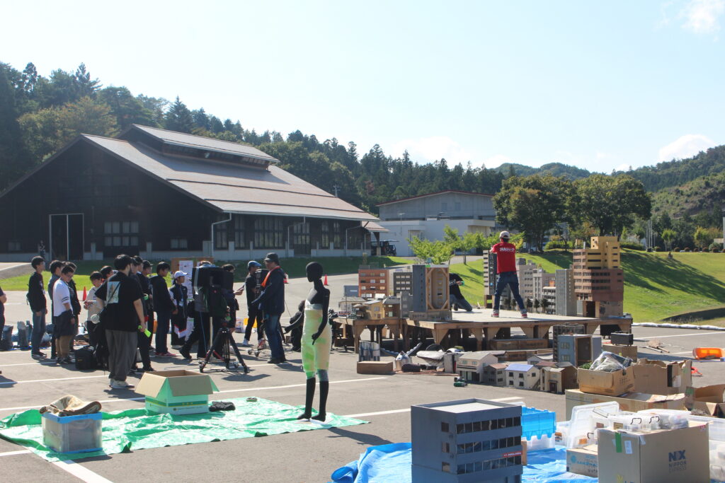 多数の参加者が野外に並べた建物模型や撮影機材の周囲で特撮セットの準備作業を進める様子の写真