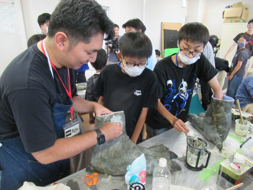 屋内の作業場で、男性が二人の少年に教えながら、大きな足の形をした模型の表面に白い塗料のようなものを塗りに取り組んでいる様子の写真