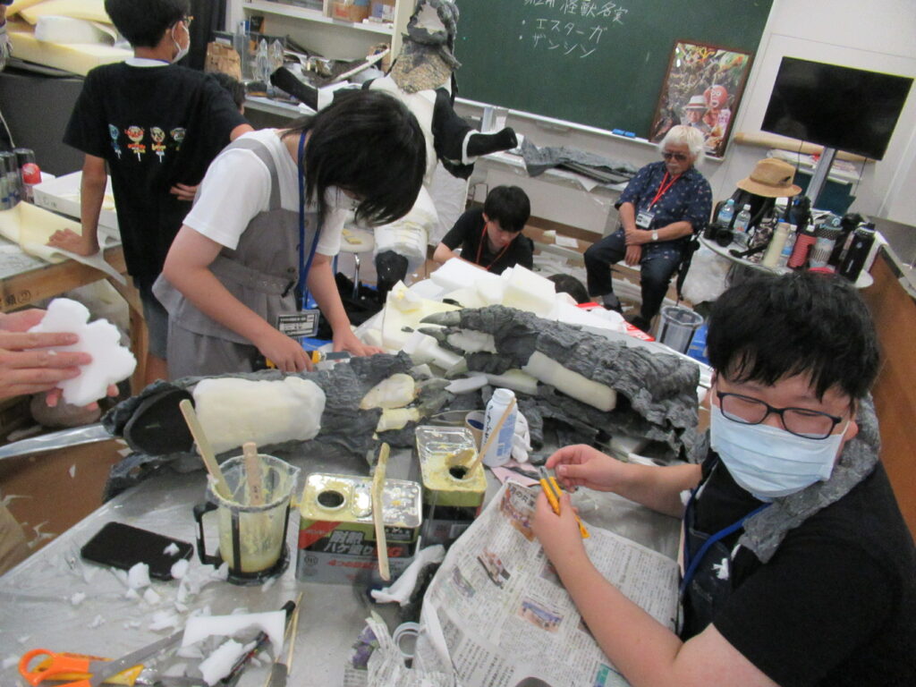 屋内の作業場で、散乱する白いスポンジ素材や接着剤の缶に囲まれながら、参加者たちが大きな怪獣の手のような模型に貼り付けたりしている制作風景の写真