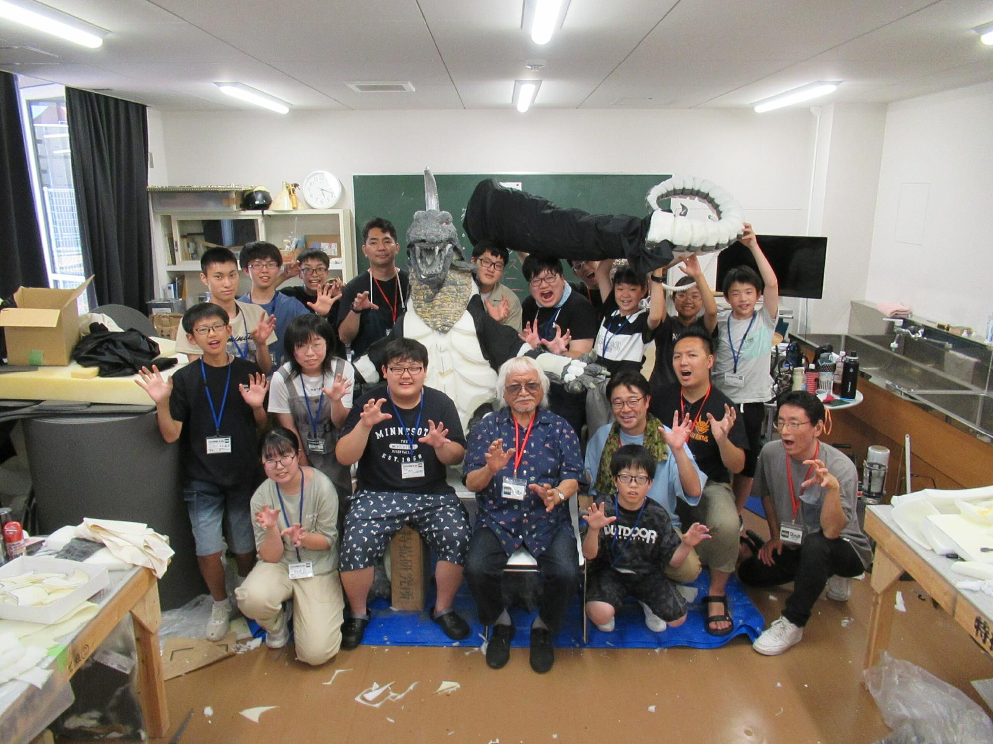 室内で、手作りの怪獣の着ぐるみや大きな尻尾の模型を囲み、子供から大人まで複数の男女が怪獣のポーズを真似て笑っている集合写真