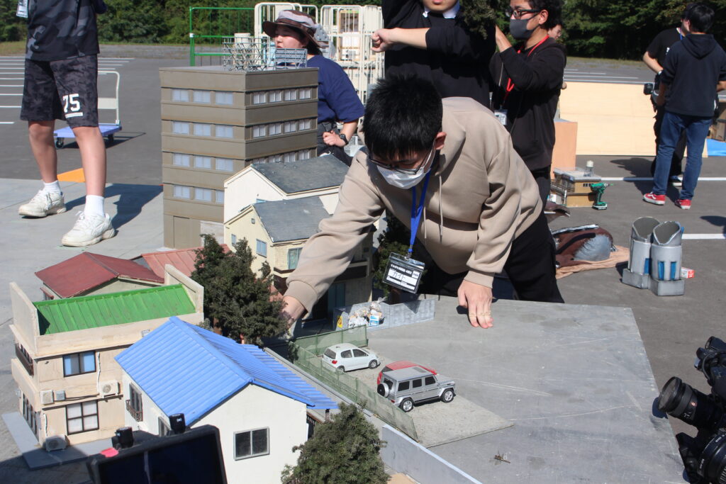 ミニチュア都市模型の建物や車の位置を丁寧に整えながら手元で配置作業を進める男性の様子の写真