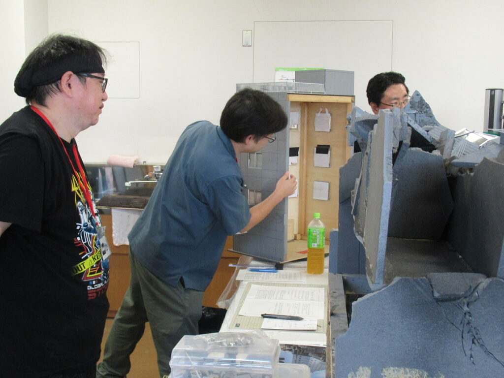 屋内の作業場で、男性たちがビルのミニチュアのセットを慎重に動かしている様子の写真