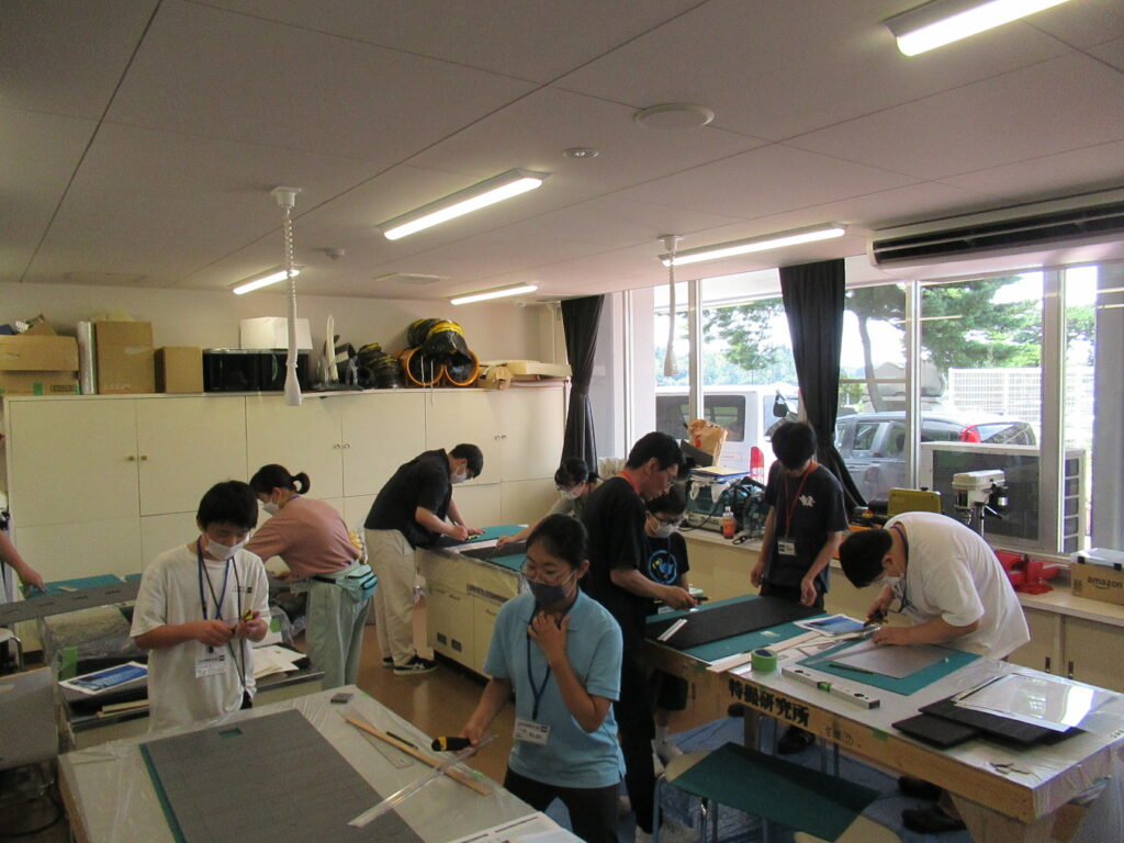 屋内の作業場で複数の男女が机に向かい、カッターや定規を使用してミニチュア制作のための素材を加工したりしている様子の写真