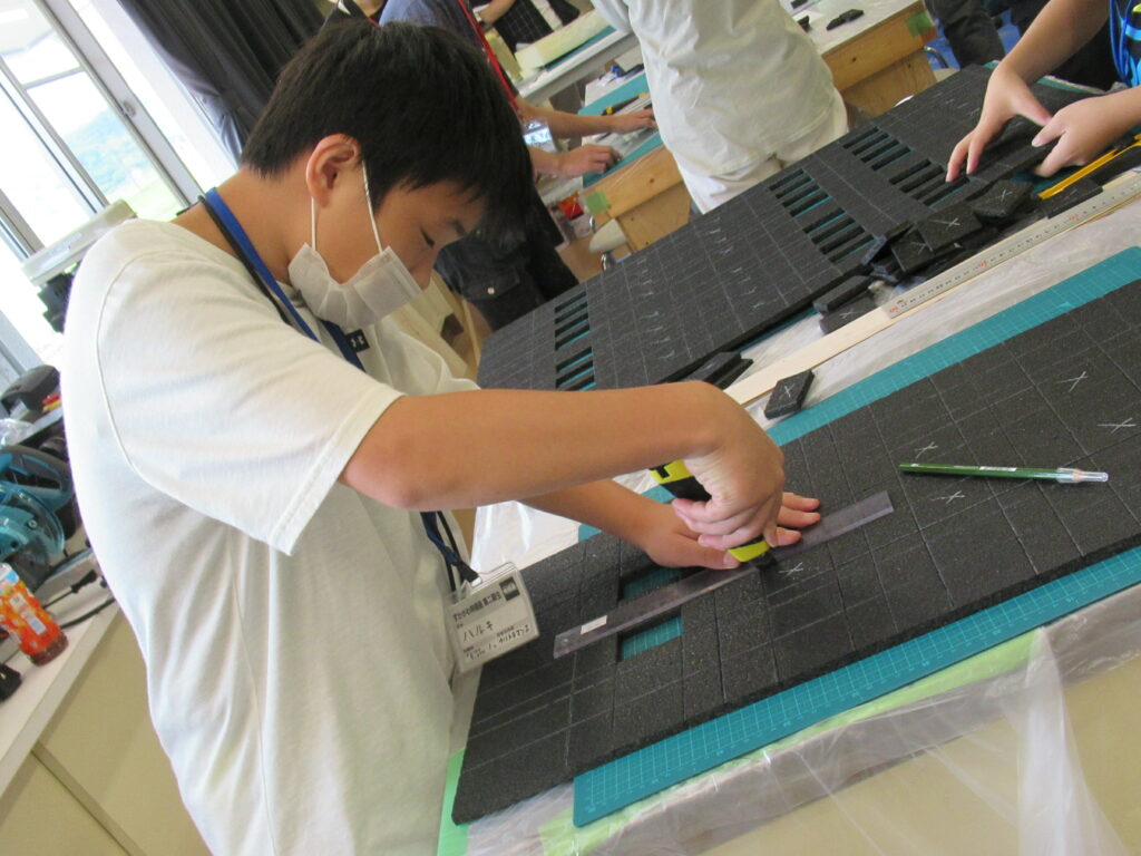 屋内の作業場で少年が、定規とカッターを使って黒いボードを四角に切っている写真