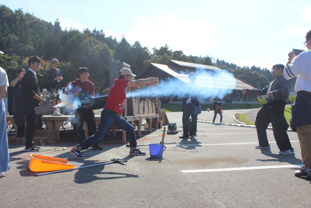 赤い上着の人物が大きく踏み込みながら装置から青い煙を放射し周囲のスタッフが撮影準備を進めている様子の写真