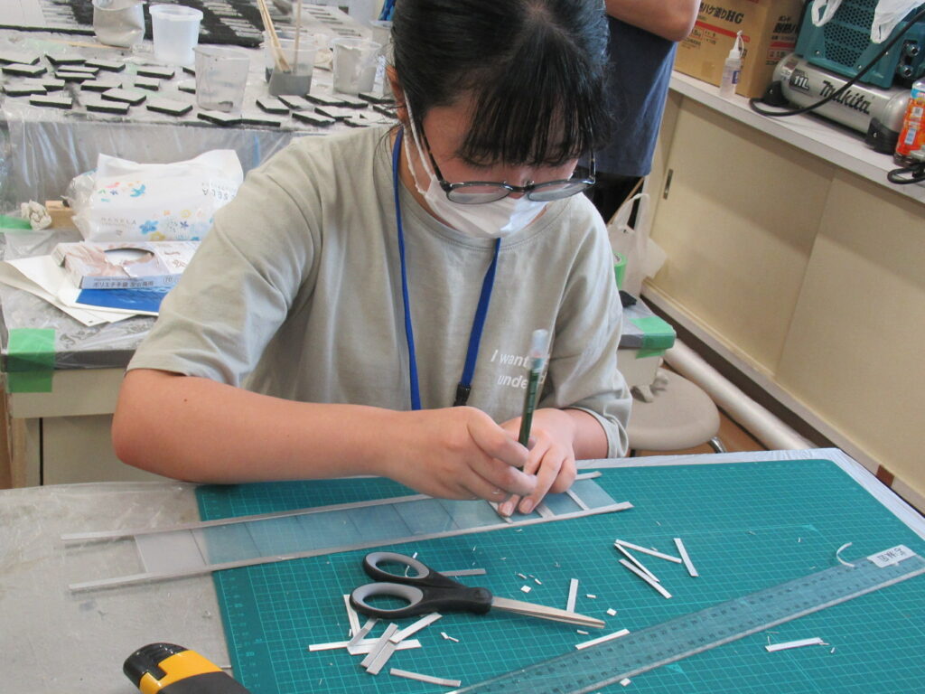 屋内の作業場で、女性がカッティングマットの上で細長い素材を慎重に合わせている様子の写真