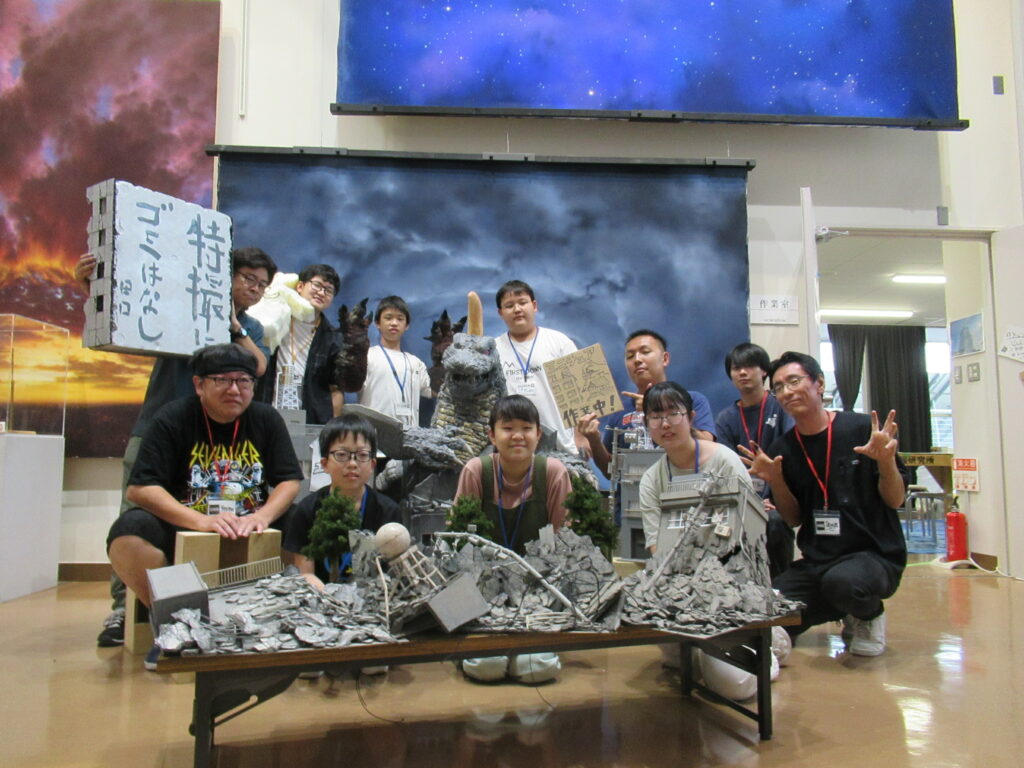 星空や雷雲の背景セットを背に、怪獣模型や破壊されたビルのミニチュアを囲んで、自作の看板や小道具を手にした塾生と講師の集合写真