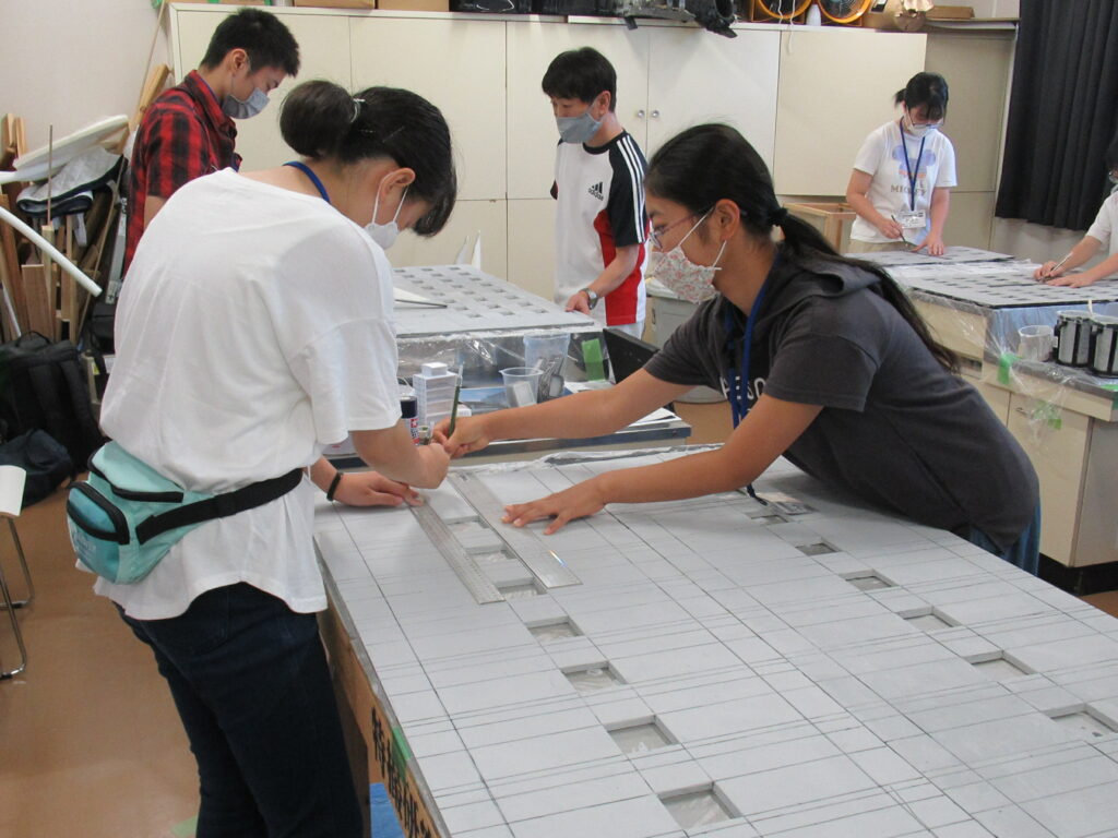 屋内の作業場で、数名の男女が大きな作業机を囲み、長い定規と鉛筆を使って薄い灰色のボードにビルの窓枠のような線を描いている様子の写真