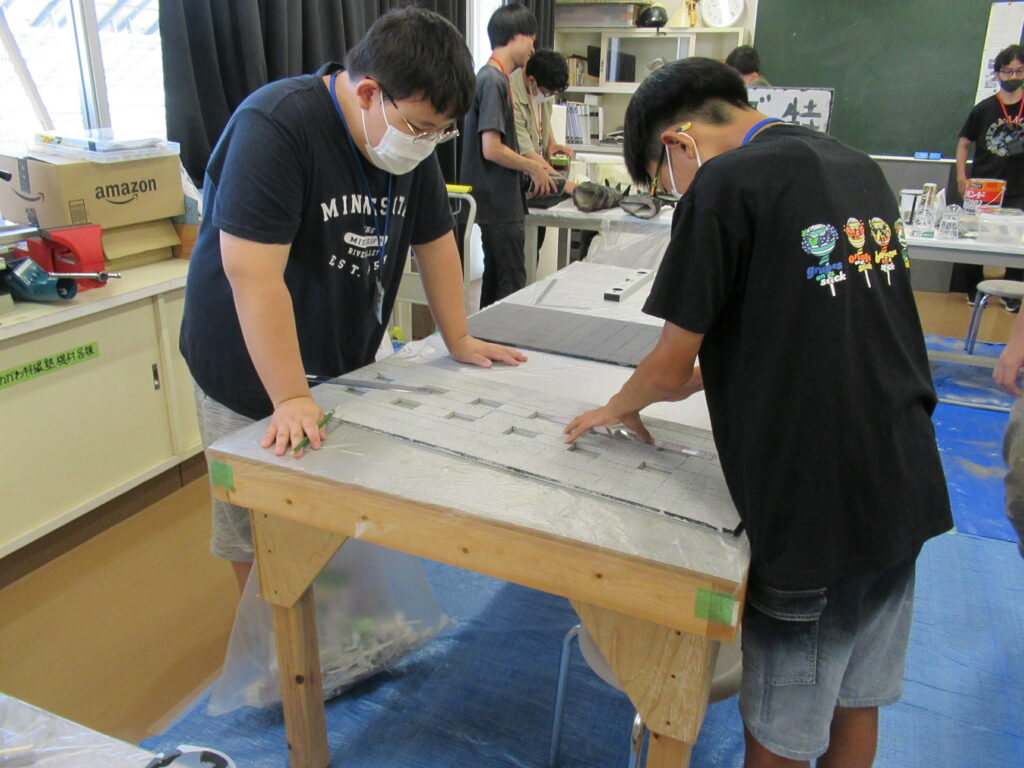 屋内の作業場で、2人の少年が木製の作業机に向かい、長い定規を用いて灰色のボードに窓枠のような線を引いたりしている様子の写真