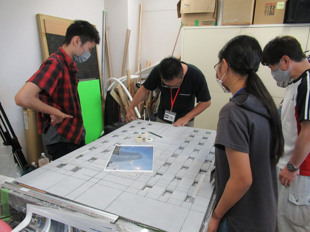 屋内の作業場で、4名の男女が大きな作業机を囲み、参考写真を見ながら灰色の巨大なビルミニチュアの表面に細かな窓枠のパーツを慎重に配置して構成を確認している様子の写真