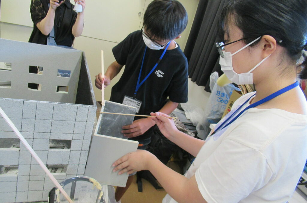 屋内の作業場で、少年と少女が、細い棒を使いながら灰色のビル型ミニチュアのパーツを持ち作業を進めている写真