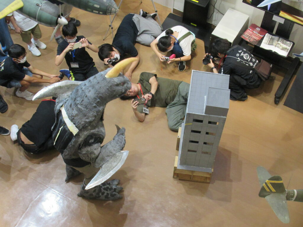 床に置かれた怪獣模とビルミニチュアに対し、塾生たちが地面に寝そべったり身をかがめたりして、スマートフォンやカメラで写真を写している写真