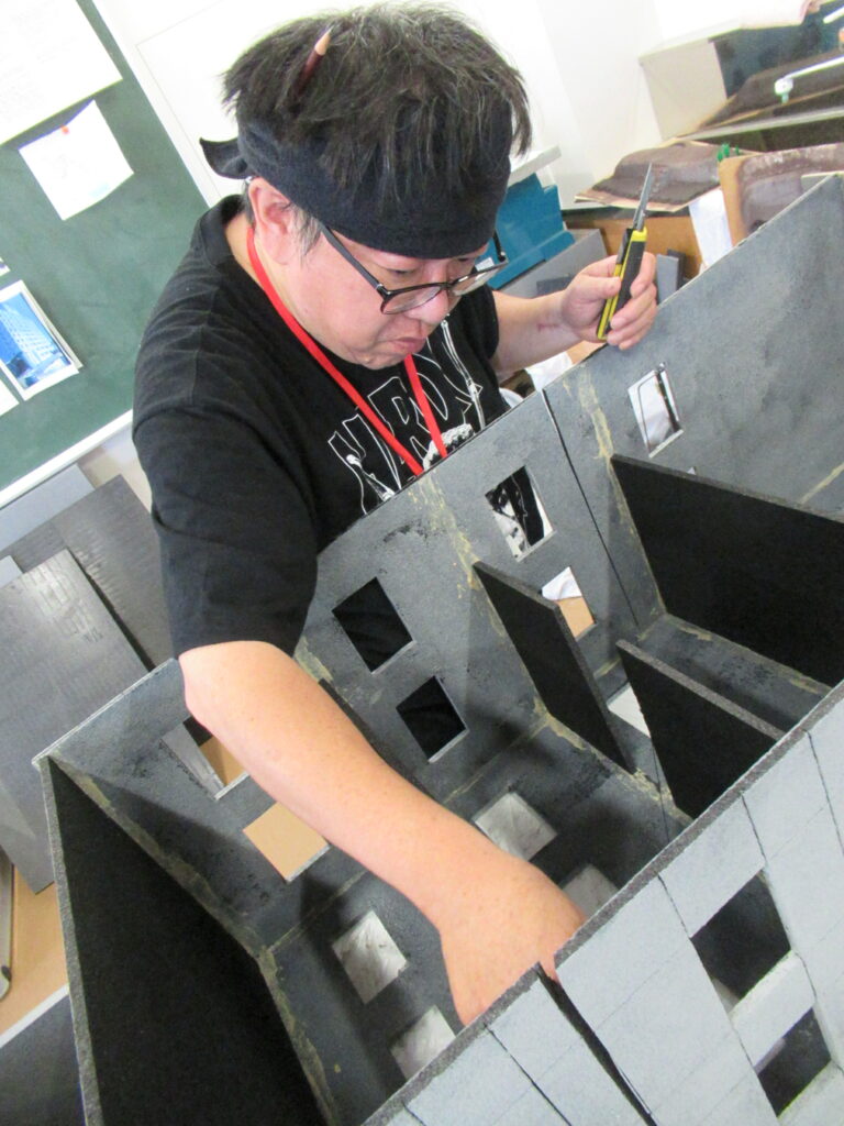 男性が手にカッターを持ち、巨大なビルミニチュアの内部構造を真剣な表情で調整している様子の写真