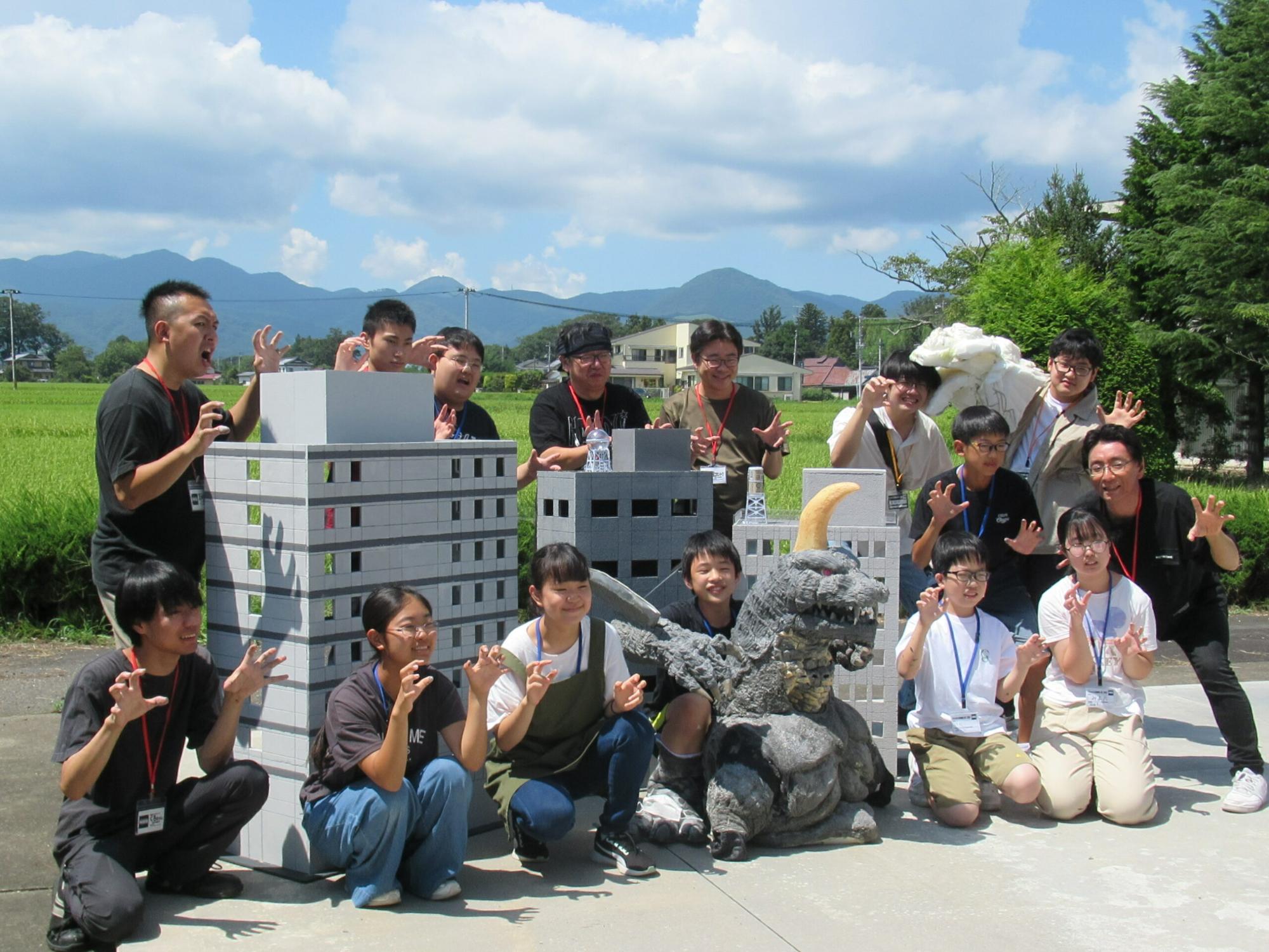 青空の下、田園風景を背景に怪獣やビルのミニチュアと一緒にが怪獣のようなポーズをした塾生と講師の集合写真