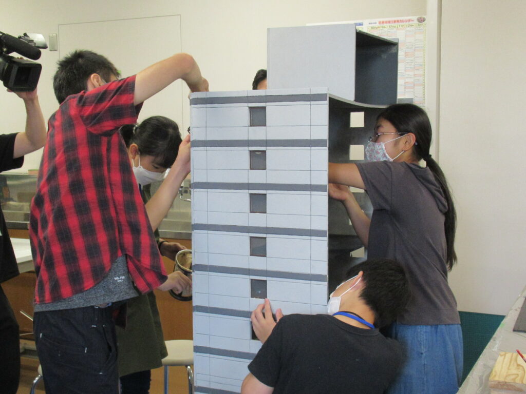 屋内の作業場で、数名の男女が大きな机を囲み、自作のビルミニチュアに接着剤を塗ったりパーツを組み上げている写真