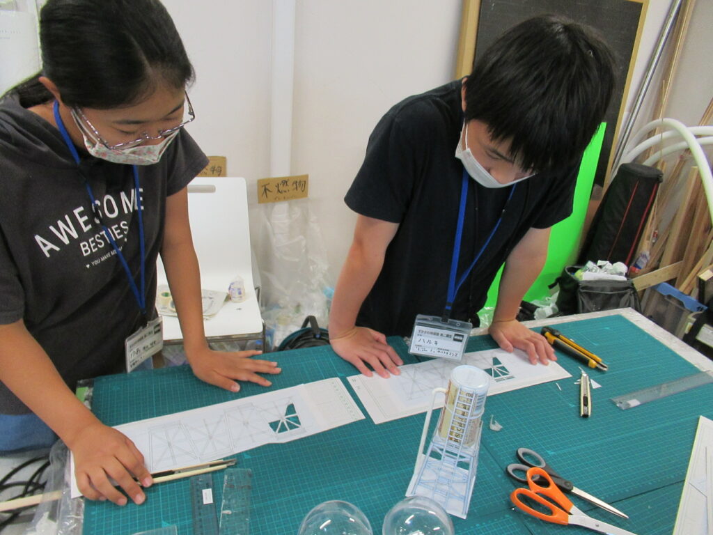 屋内の作業場で男女の塾生たちが、机の上に広げられた切った図面を確認している様子の写真