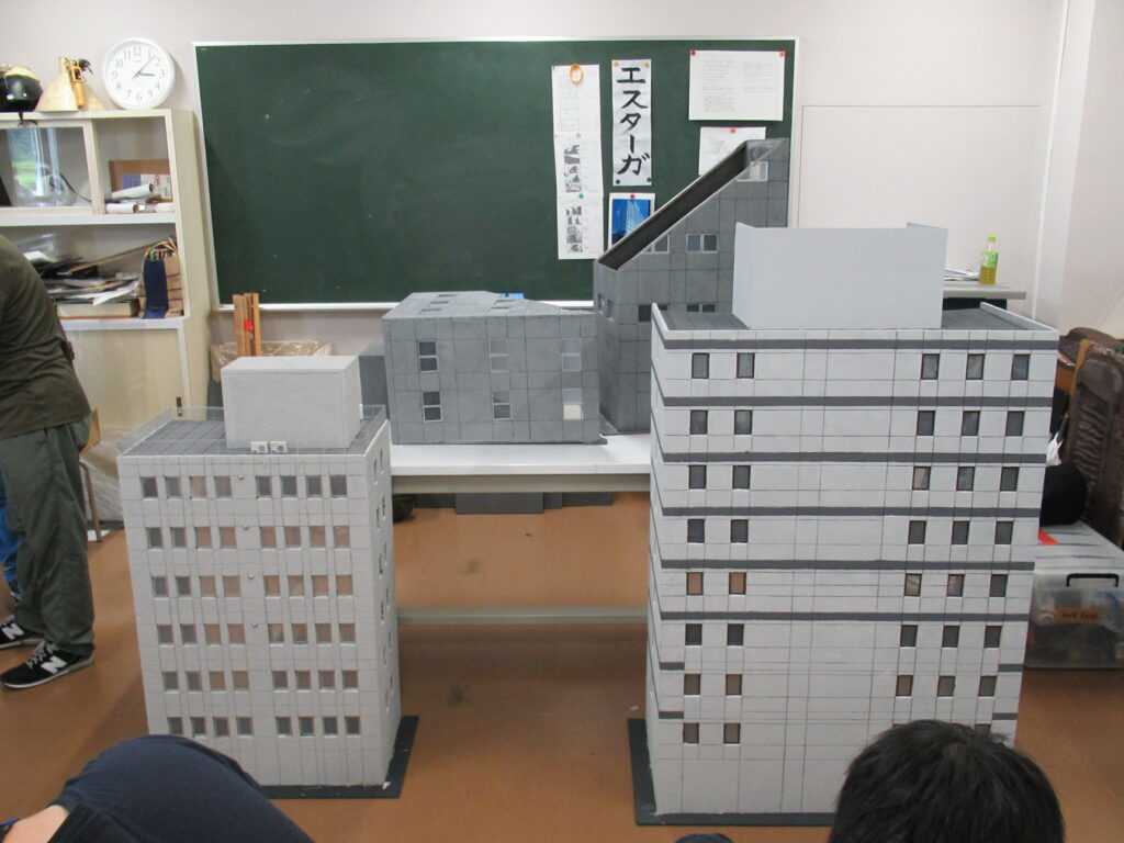 屋内の作業場で、さまざまな形状をした複数のビル型ミニチュア模型が机の上や床に並べられている写真