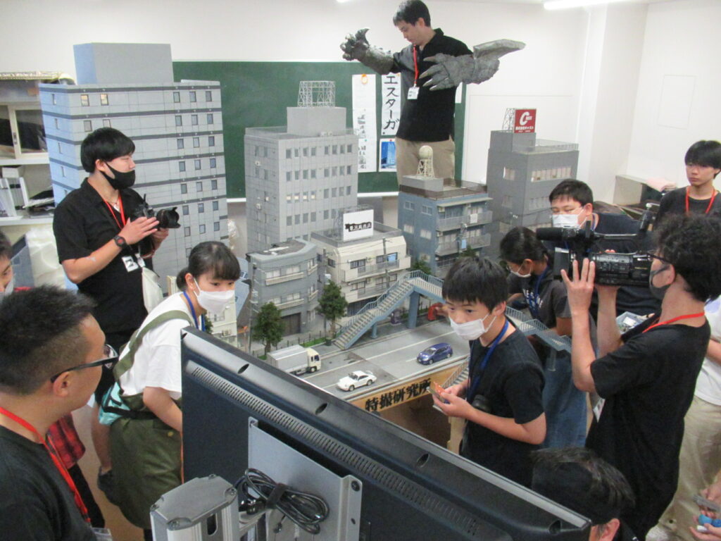屋内に道路や歩道橋が再現されたミニチュアセットの横に怪獣の腕を装着したスタッフが立ち、ビデオカメラを構える人やモニターを確認する塾生たち写真