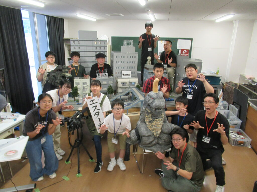 屋内の作業場で、完成したミニチュアセットや怪獣模型を囲み、塾生たちが怪獣のようなポーズをとっている集合写真