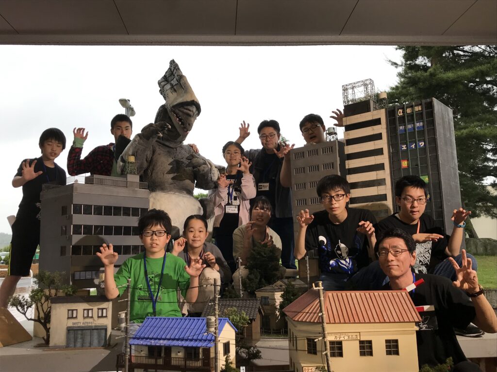 高層ビルや民家が立ち並ぶミニチュアセット、巨大な怪獣とすかがわ特撮塾生と講師がいっしょに並び、全員が怪獣を模したポーズをしている写真